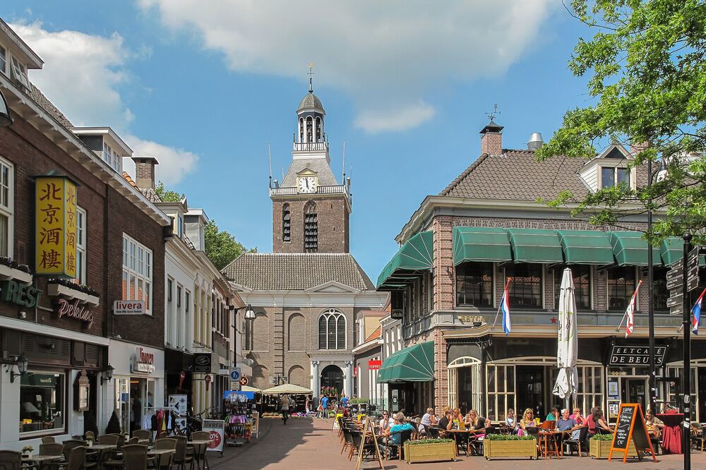 De Meppeler toren rijst op boven een zonnige winkelstraat in het centrum van Meppel, met terrassen, marktkraampjes en wandelend publiek